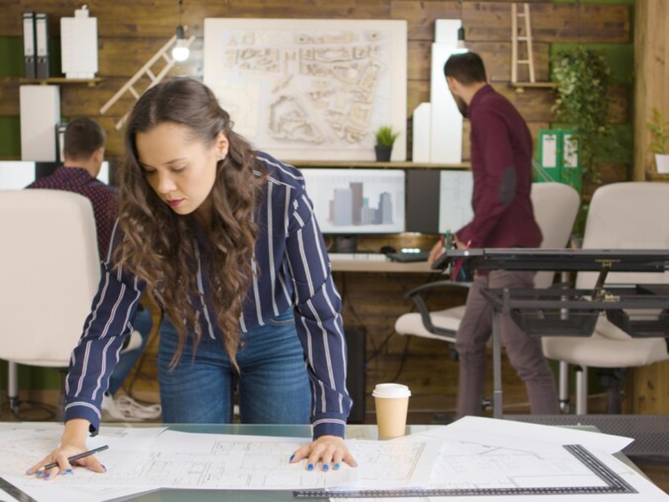 Business woman standing at her desk, leaning over it and reviewing plans and papers on her desk.