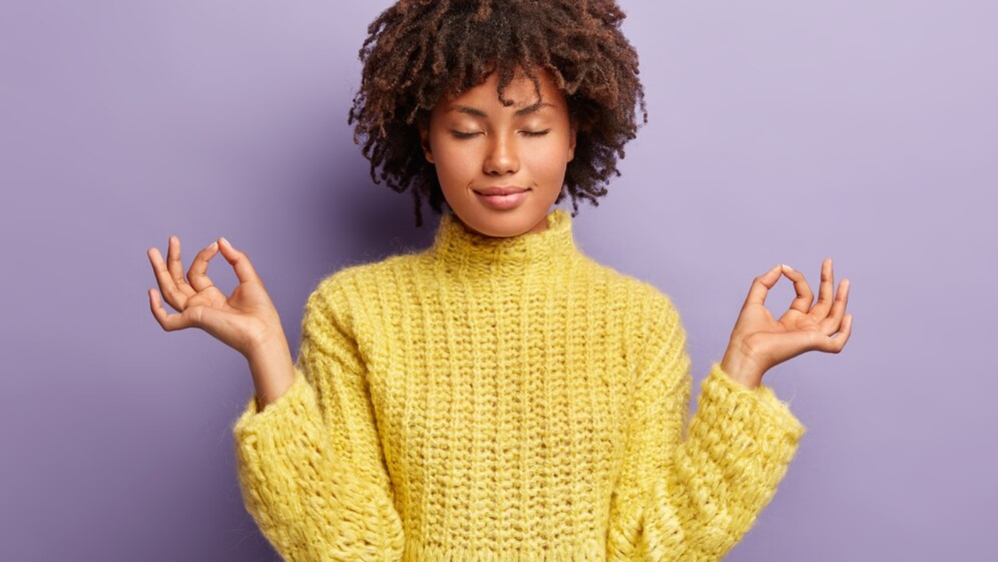 young woman with loose curls afro haircut wearing a yellow sweater. Her eyes are closed and she seems to be contently meditating.