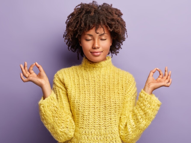 young woman with loose curls afro haircut wearing a yellow sweater. Her eyes are closed and she seems to be contently meditating.
